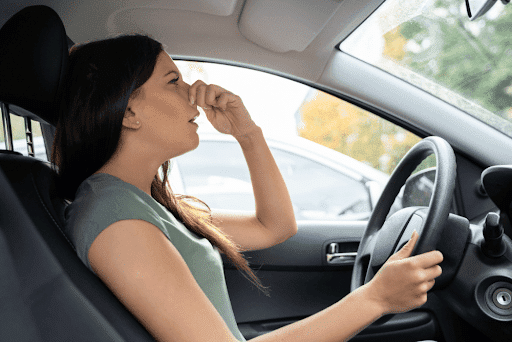 Weird car smells and their causes in Marion, IL with Hale’s Automotive. Image of young woman driving her car and plugging her nose with her left hand due to a weird car smell.